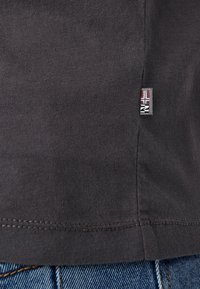 Tummanharmaa puuvillainen t-paita, jossa on sileä pinta ja pieni neliömäinen, punavalkoisilla yksityiskohdilla varustettu etiketti ommeltuna helmaan.