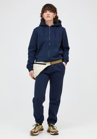 Felpa con zip color navy e tasche frontali, abbinata a pantaloni della tuta navy. Marsupio bianco trapuntato in vita e scarpe da ginnastica beige con dettagli colorati.