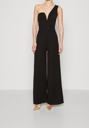 Schwarzer One-Shoulder-Jumpsuit mit tiefem V-Ausschnitt und weitem Bein, hergestellt aus einem glatten Stoff. Kombiniert mit transparenten Absatzschuhen.