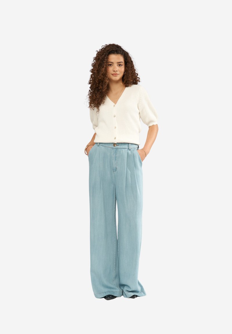Femme aux cheveux bouclés portant un haut blanc à manches courtes boutonné et un pantalon large et ample bleu clair, debout les mains dans les poches.
