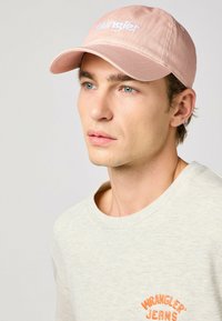 Gorra de béisbol color rosa claro con el logo "Wrangler" bordado en blanco. Tejido suave, visera curva y correa trasera ajustable. El modelo lleva puesta una sudadera de color beige claro.