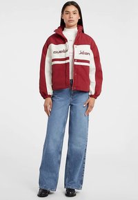 Rød og cremefarvet oversized jakke med elastiske manchetter, med logo tekst foran, kombineret med blå bukser med vide ben og sorte støvler.