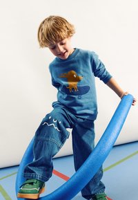 Sweatshirt bleu clair avec un graphisme de chien marron, associé à un jean bleu avec des motifs de vagues blanches sur les genoux et des chaussures vertes.