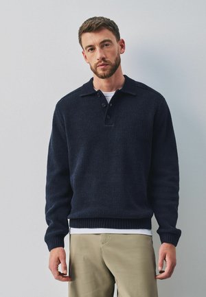 Mann trägt einen dunkelblauen Strickpullover über einem weißen Hemd und beigen Hosen, steht vor einem schlichten hellgrauen Hintergrund.