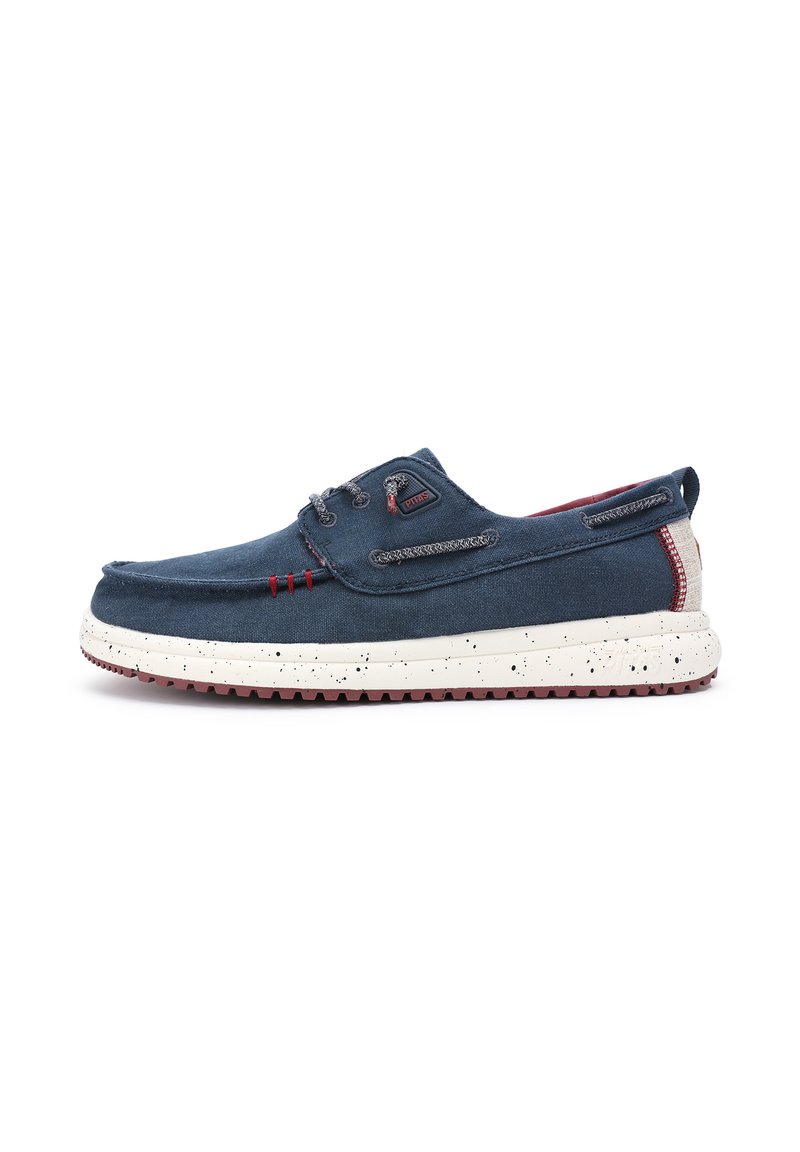 Zapato de barco de lona en azul marino con una parte superior texturizada, suela blanca moteada, acentos rojos y costuras decorativas. Presenta un diseño sin cordones.