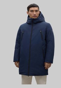 Parka aislante de color azul marino con capucha, que presenta cremalleras negras en la parte delantera y en el pecho. Textura suave, mangas largas y corte recto.