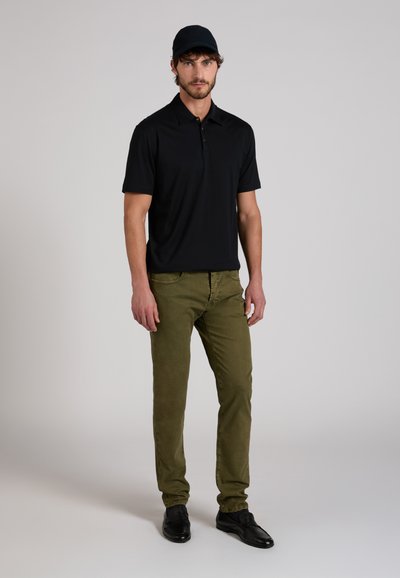 Man die staat en een zwarte pet draagt, een zwart poloshirt met korte mouwen, olijfgroene broek en zwarte nette schoenen, tegen een neutrale achtergrond.