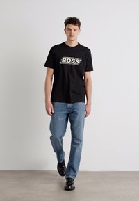 T-shirt en coton noir avec le logo "BOSS" en blanc, associé à un jean bleu clair et des chaussures noires, arborant un design simple et droit.