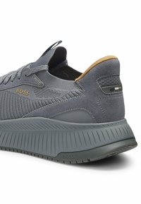 Gray athletic shoe featuring a textured fabric upper, suede accents, and a ridged rubber sole. Gold branding detail on the side.