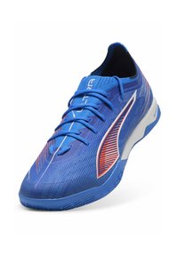 Niebieskie buty sportowe z teksturowaną siateczkową cholewką, kontrastującymi czerwonymi akcentami i białą podeszwą środkową. Zawierają sznurówki i zaokrąglony kształt czubka.