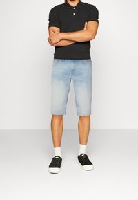 Camisa polo negra con tres botones, pantalones cortos de mezclilla azul claro hasta las rodillas, calcetines blancos y zapatillas negras con suelas blancas.