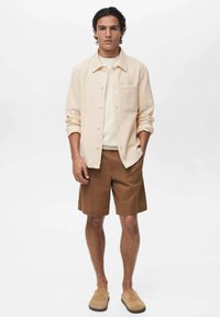 Light beige button-up shirt over a white sweater, paired with brown shorts and tan slippers. Simple design, textured fabric, and neutral colors.