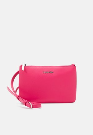 Jasnoróżowa saszetka na nadgarstek Calvin Klein z zamkiem błyskawicznym i regulowanym paskiem, wykonana z gładkiego materiału o fakturze skóry.