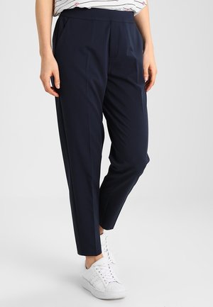 Femme portant un pantalon fuselé bleu marine foncé, des baskets blanches et une chemise blanche à rayures avec de petits motifs de cœurs, debout devant un fond uni.