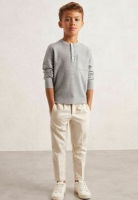 Camiseta henley gris con canalé y un bolsillo en el pecho, combinada con pantalones slim-fit color crema y zapatillas blancas, sobre un fondo neutro.
