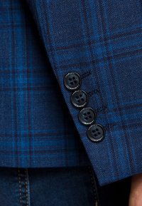 Niebieska marynarka w kratę z rękawem z teksturowanej tkaniny, czterema czarnymi guzikami z logo oraz precyzyjnymi detalami szwu w pobliżu mankietu.