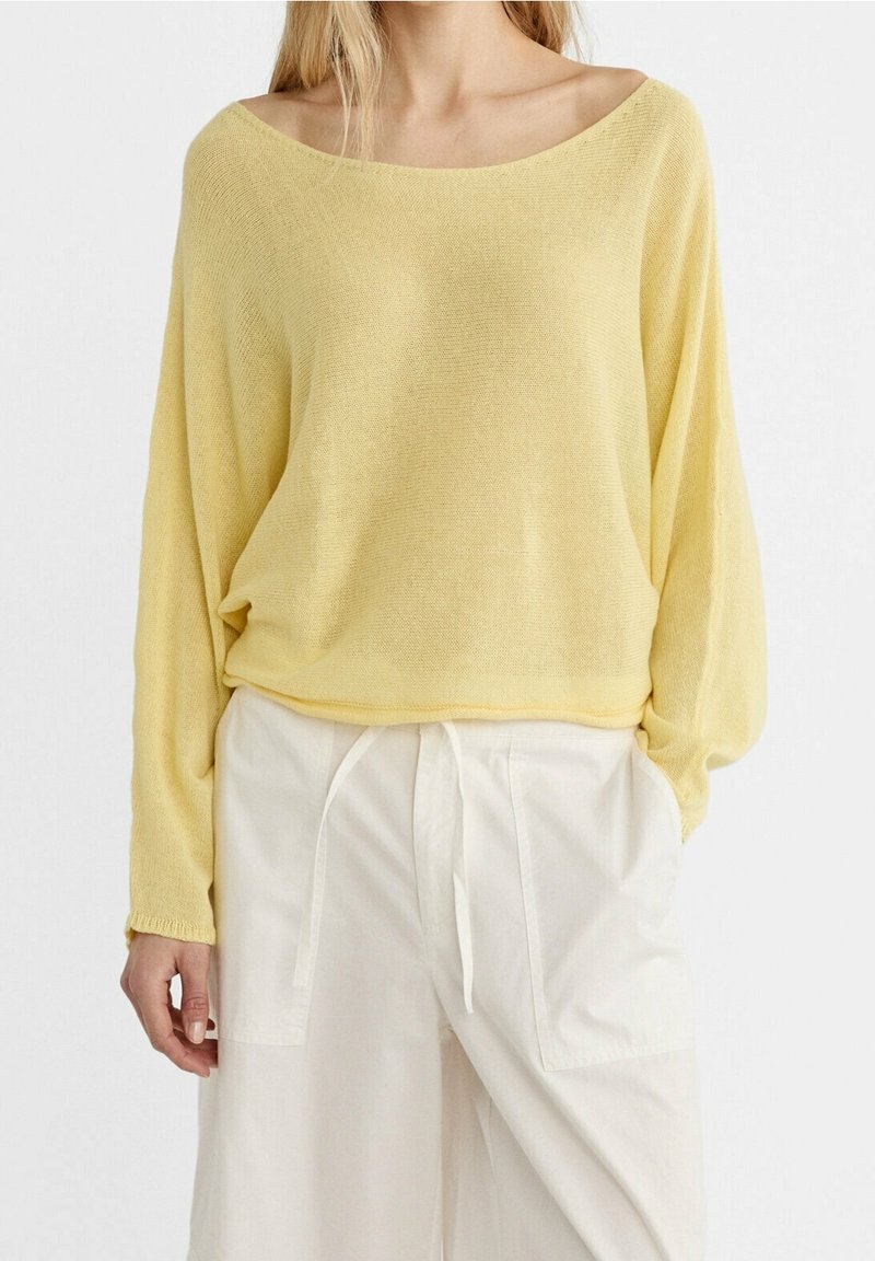 Femme portant un pull ample à manches longues de couleur jaune clair avec un col large, ainsi qu'un pantalon blanc taille haute avec poches avant et cordon de serrage.