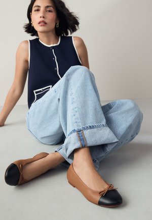 Mujer con cabello corto y oscuro sentada con las piernas cruzadas, vistiendo una camiseta sin mangas azul marino, vaqueros claros y zapatos planos color beige con puntera negra.