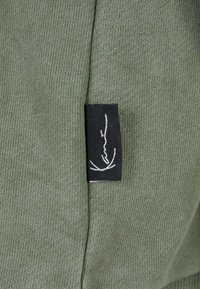 Gros plan d’un tissu tricoté vert avec une étiquette rectangulaire noire cousue dans la couture, portant un texte en écriture cursive blanche.