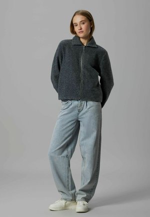 Fiatal nő áll, kezét a háta mögött tartja, szürke cipzáras dzsekit, világoskék farmert és fehér tornacipőt visel, szürke háttér előtt.