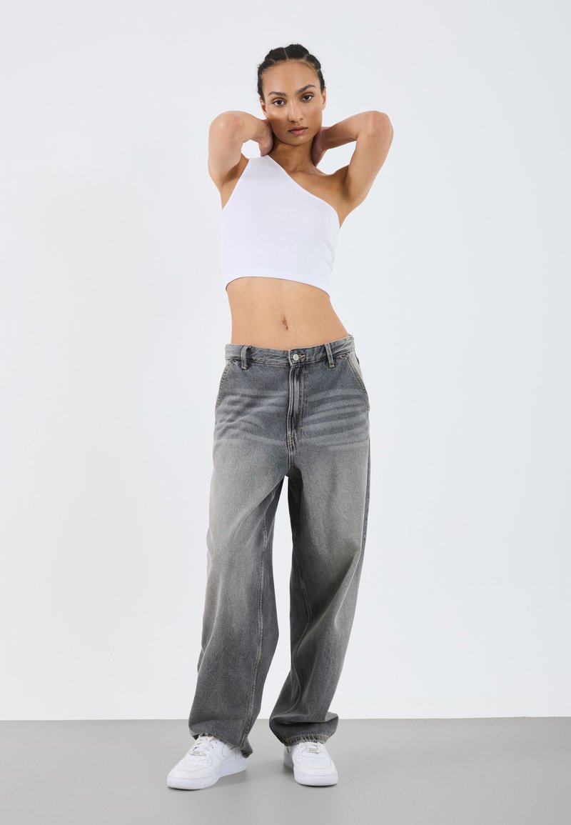 Jeans anchos de corte recto en gris, desgastados y con cintura alta, combinados con un top corto asimétrico blanco y zapatillas blancas sobre un suelo gris.