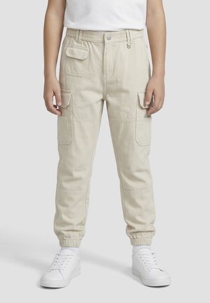 Kind trägt beige Cargohosen mit elastischen Bündchen und Klappentaschen, weiße Sneaker und ein weißes T-Shirt, steht vor einem einfarbigen Hintergrund.