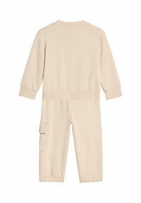Beżowy, długorękawowy kombinezon z elastycznymi mankietami i pasem w talii. Posiada boczną kieszeń cargo oraz gładką fakturę materiału. Widok z tyłu.