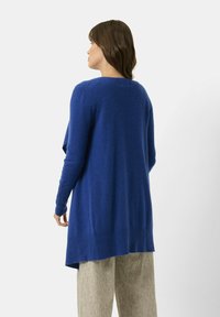 Pitkä sininen neulejakku, jossa on ribbireunat hihoissa ja helmassa. Pehmeä rakenne ja löysä malli, jonka hi-lo helma ulottuu vaaleanharmaiden housujen ylle.