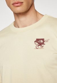 Beige bomulls t-skjorte med et lekent, trykt logo i brunt som skildrer en karakter ved siden av en datamaskin med teksten "THE NORTH FACE".