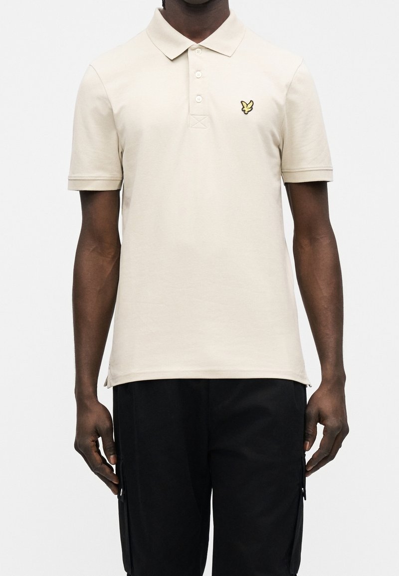 Homme portant un polo beige clair à manches courtes avec un petit emblème jaune d’oiseau sur la poitrine et un pantalon noir.
