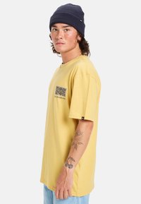 Žluté bavlněné tričko s grafickým potiskem na levé straně hrudi, spárované s džínovými šortkami. Model má na sobě tmavou beanie a tetování na jedné ruce.