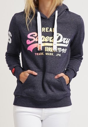 Ciemnoniebieska bluza z kapturem z białymi troczkami, różowo-żółte logo "SuperDry", przednie kieszenie, noszona przez osobę w białych spodniach z rękami w kieszeniach.