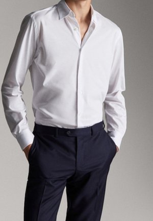 Homme portant une chemise blanche ajustée à manches longues boutonnée, rentrée dans un pantalon bleu marine foncé sur mesure, les mains dans les poches.