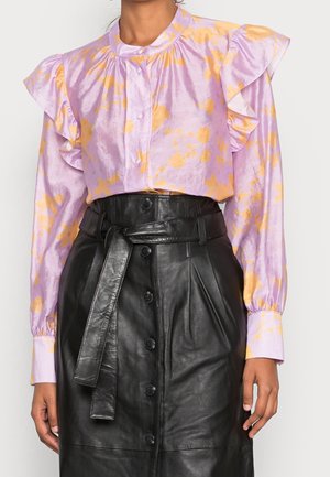 Femme portant un chemisier violet à imprimé floral jaune avec des épaules à volants, associé à une jupe en cuir noire taille haute boutonnée avec ceinture.