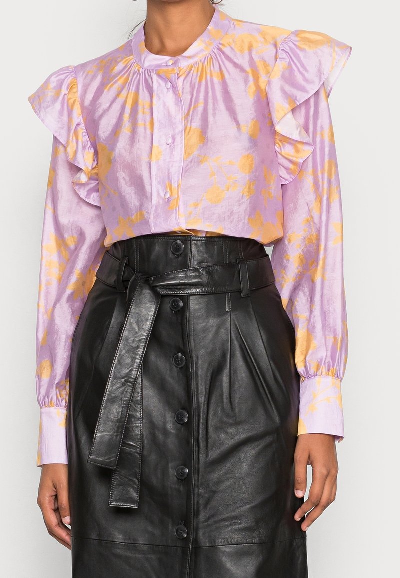 Femme portant un chemisier violet à imprimé floral jaune avec des épaules à volants, associé à une jupe en cuir noire taille haute boutonnée avec ceinture.