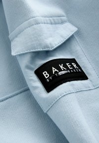 Hele sinine kangas tekstuurse pinnaga, millel on must silt tekstiga "BAKER BY TED BAKER." Sisaldab ka voltis krae detaili.