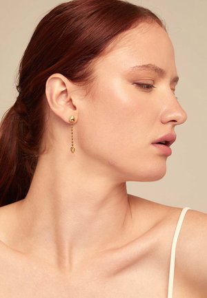 Los pendientes de oro colgantes presentan un acento en forma de lágrima, una parte superior esférica lisa y un gancho de cadena, mostrados en el perfil de un modelo.