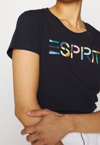 Černé bavlněné tričko s krátkými rukávy, zdobené barevným logem "ESPRIT" v různých vzorech. Design s kulatým výstřihem, hladká textura.