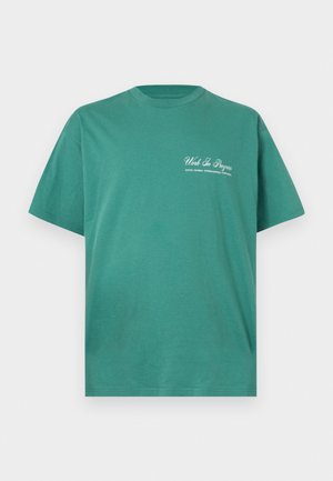 Zielony T-shirt z krótkim rękawem i okrągłym dekoltem z białym napisem „Work In Progress” oraz mniejszym tekstem poniżej, umieszczonym na lewej górnej części klatki piersiowej.