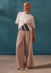 Λευκό κοντομάνικο μπλουζάκι με μαύρο γραφικό σχέδιο στο μπροστινό μέρος, σε συνδυασμό με χαλαρές μπεζ παντελόνες με ελαστική μέση και λογότυπο.