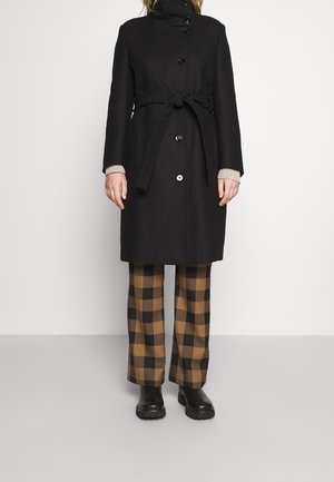 Personne portant un manteau noir ceinturé, un pantalon à carreaux marron et noir, ainsi que des bottes noires, debout sur un fond blanc uni.