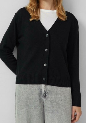 Femme portant un cardigan noir boutonné sur un haut blanc et un jean gris clair, debout avec une main dans le dos.