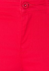 Tela de mezclilla roja con un bolsillo frontal, cierre de botón y costuras visibles. Textura suave con un acabado ligeramente brillante.