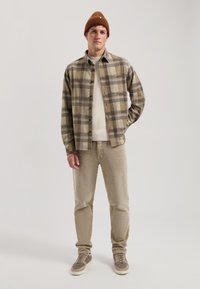 Cămașă din flanel cu carouri în nuanțe de maro și bej, pulover bej dedesubt, pantaloni bej deschis și adidași gri cu o căciulă maronie.