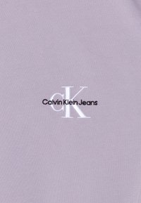 Světle fialová bavlněná tkanina s vyšitým černým logem "Calvin Klein Jeans" a bílým akcentem "cK". Hladká textura a jednoduchý design.