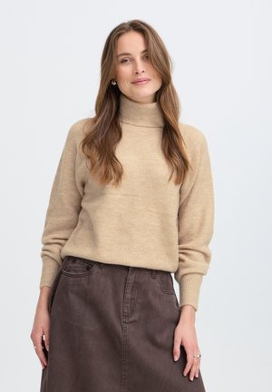 Jeune femme aux longs cheveux bruns portant un pull à col roulé beige et une jupe marron foncé, debout devant un fond uni.