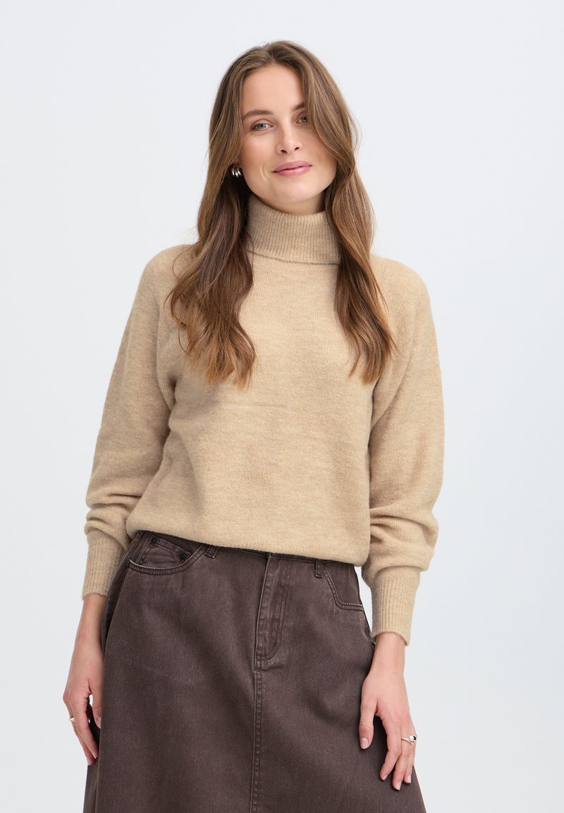 Jeune femme aux longs cheveux bruns portant un pull à col roulé beige et une jupe marron foncé, debout devant un fond uni.