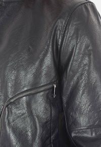 Giacca in pelle nera con superficie texturizzata, tasca con zip sul lato e un design elegante e aderente con dettagli di cucitura minimi.