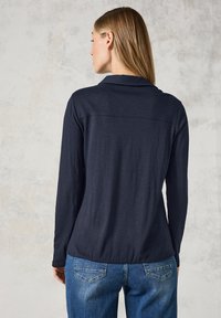 Maglietta a maniche lunghe di un blu navy scuro con colletto e vestibilità rilassata; presenta una texture liscia e dettagli delle cuciture lungo la schiena.