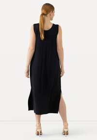 Abito lungo nero senza maniche con spacchi laterali, scollatura arrotondata e una silhouette fluida. Indossato con scarpe con tacco di colore chiaro.